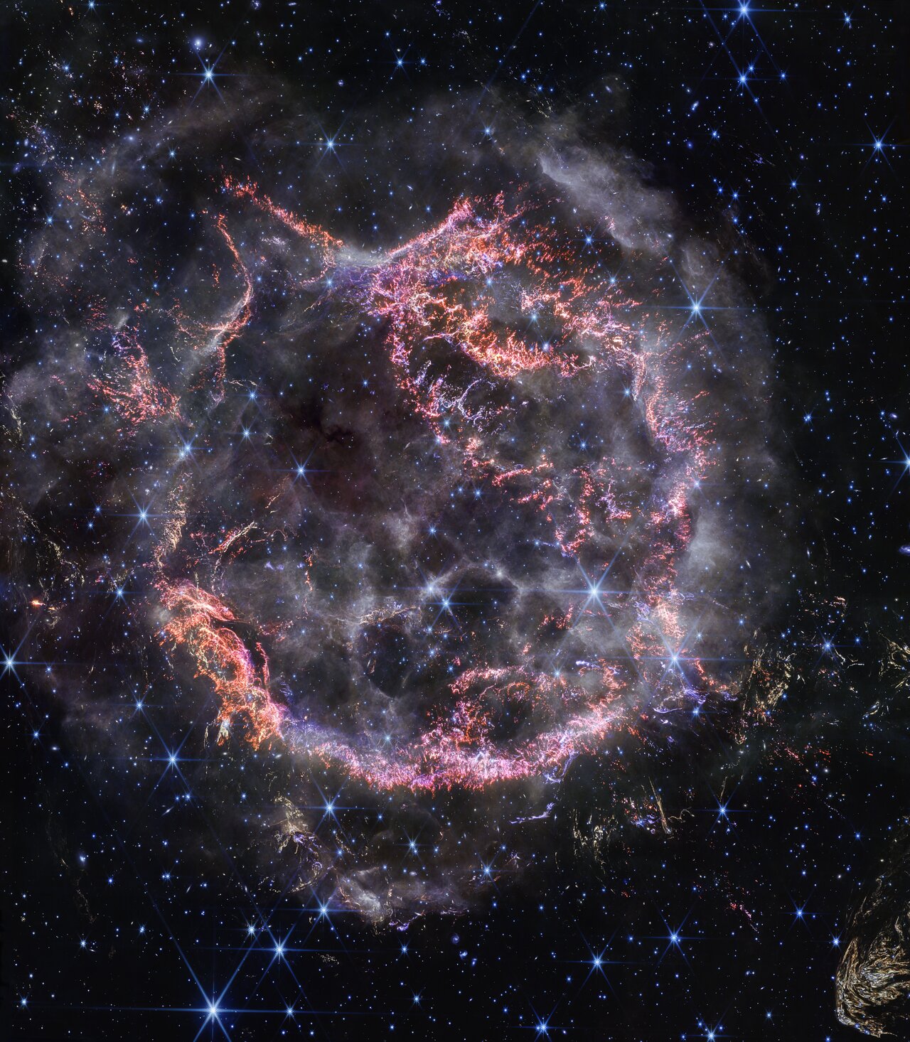 Cassiopeia A: a roughly round cloud of pink and orange filaments and knots, with smoky outer edges